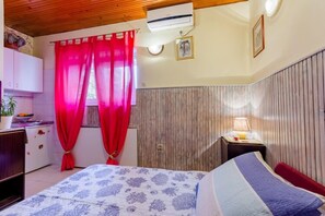 Een slaapkamer met een bed, nachtkastje met lamp, een houten garderobe en een raam met rode gordijnen.
