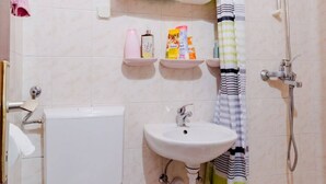Un baño con inodoro, lavamanos y ducha. Hay repisas con artículos de tocador sobre el lavamanos.