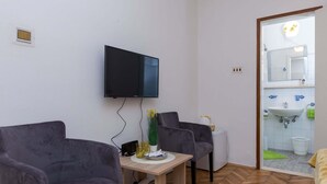 A modern room with two grey armchairs, a wooden table, a flat-screen TV, and a bathroom visible through an open door. - Sunset Bay Room - Double Room With Terrace and Partial Sea View (Dubrovnik)