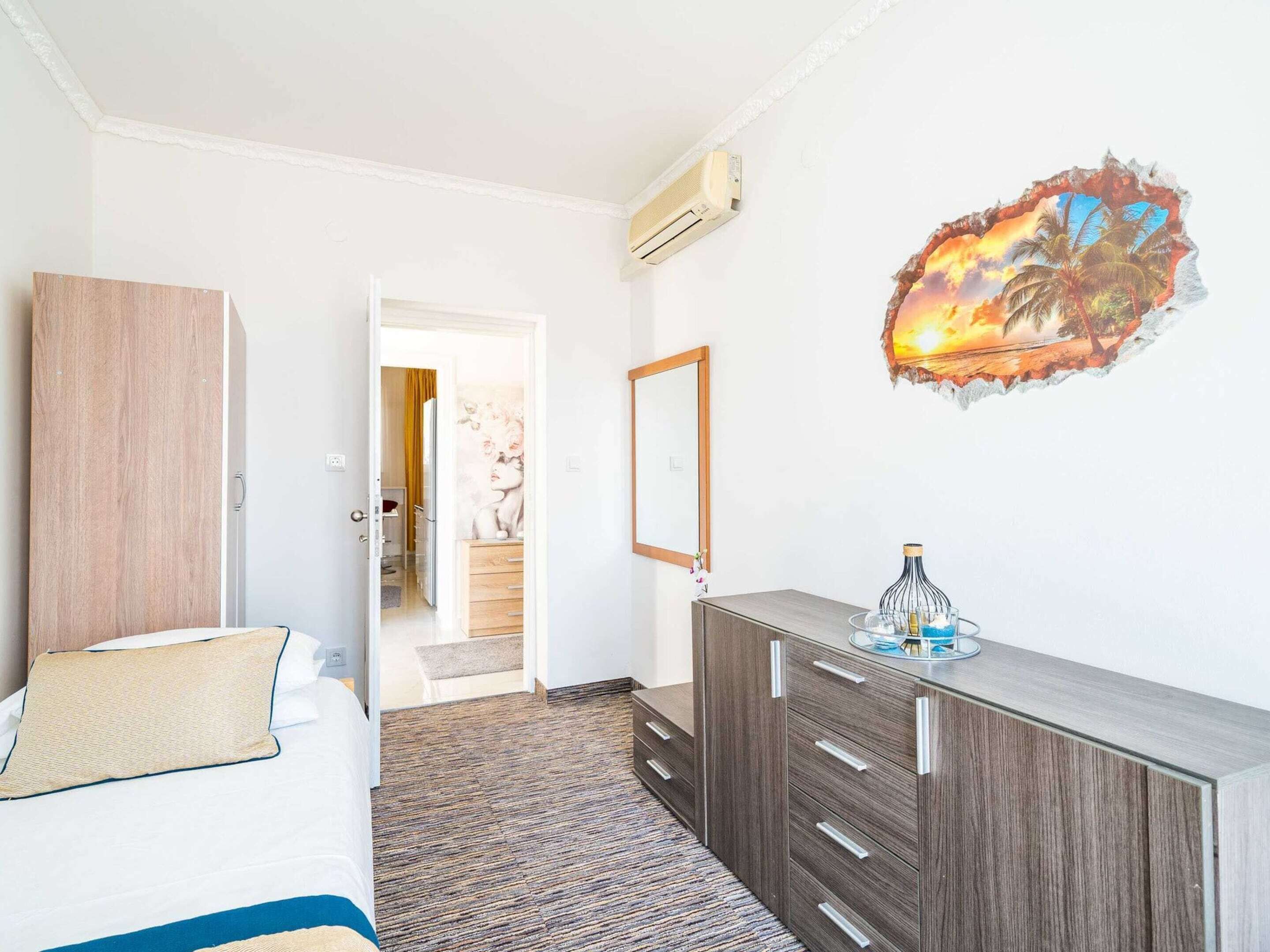 Habitación de hotel con cama, armario de madera, cómoda y una pared decorada con un mural de atardecer tropical.