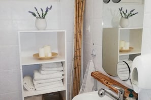 Un baño con repisas blancas que sostienen toallas y velas, un perchero de madera, un espejo y una cortina de bambú.