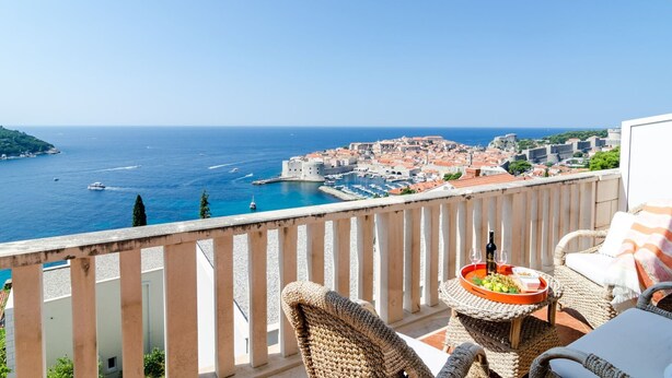 A balcony with wicker furniture, a table with a fruit bowl and bottles, and a view of a coastal town with a castle. - Apartments Bonavista - Superior Two Bedroom Apartment With Balcony and Sea View (Dubrovnik)