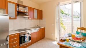 A modern kitchen with wooden cabinets, a stainless steel refrigerator, and a dishwasher. A dining table with breakfast items is set near a sliding glass door leading to a balcony. - Apartments Bonavista - Superior Two Bedroom Apartment With Balcony and Sea View (Dubrovnik)