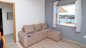 Ein Zimmer mit einem beigefarbenen Ecksofa, einem Fenster mit Vorhängen und Blick auf ein Gewässer sowie Gebäude.
