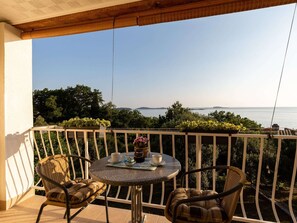 A balcony with a table and two wicker chairs, overlooking a scenic view of the sea and greenery.