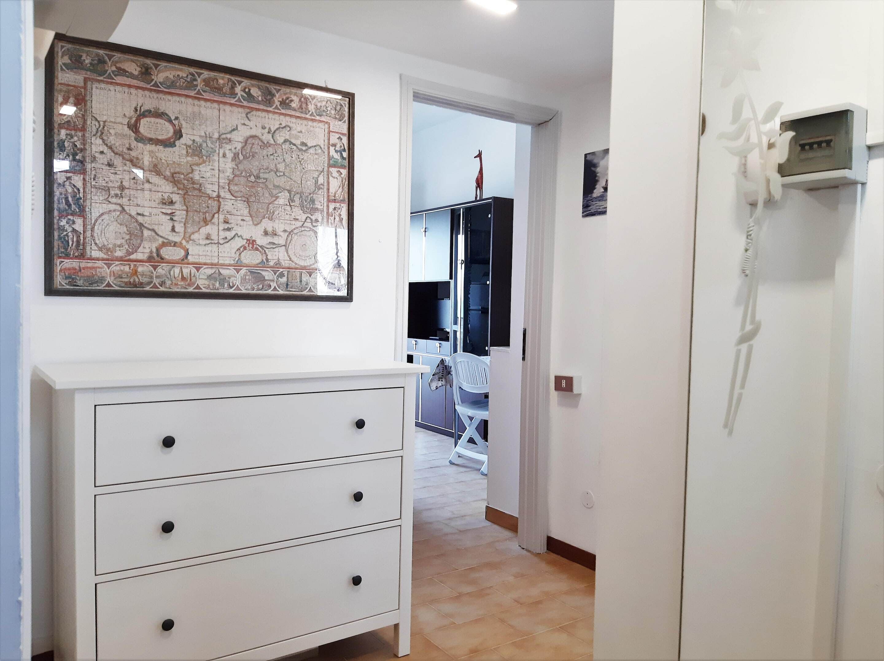 Um corredor com um aparador branco, um mapa emoldurado e uma porta que dá para um quarto com televisão e poltrona.