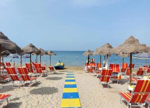 A beach with rows of red and yellow striped lounge chairs, a blue and yellow striped pathway, and thatched umbrellas. - Apartment on Marche Coast With Sea View (Mondolfo)