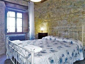 A bedroom with a stone wall, a bed with a floral bedspread, a window with shutters, and a radiator. - Belvilla by OYO Mansion in Orvieto (Orvieto)