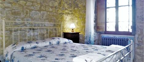 A bedroom with a stone wall, a metal bed frame, a floral bedspread, a nightstand with a lamp, and a window with a view.