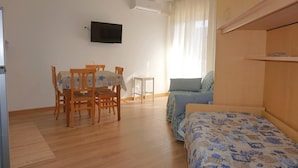 Una habitación con cama, una mesa con sillas, un televisor montado en la pared y un ventanal con cortinas.