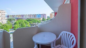 Un balcón con una mesa y silla de plástico blanco, un muro rojo y vistas a edificios coloridos y árboles.