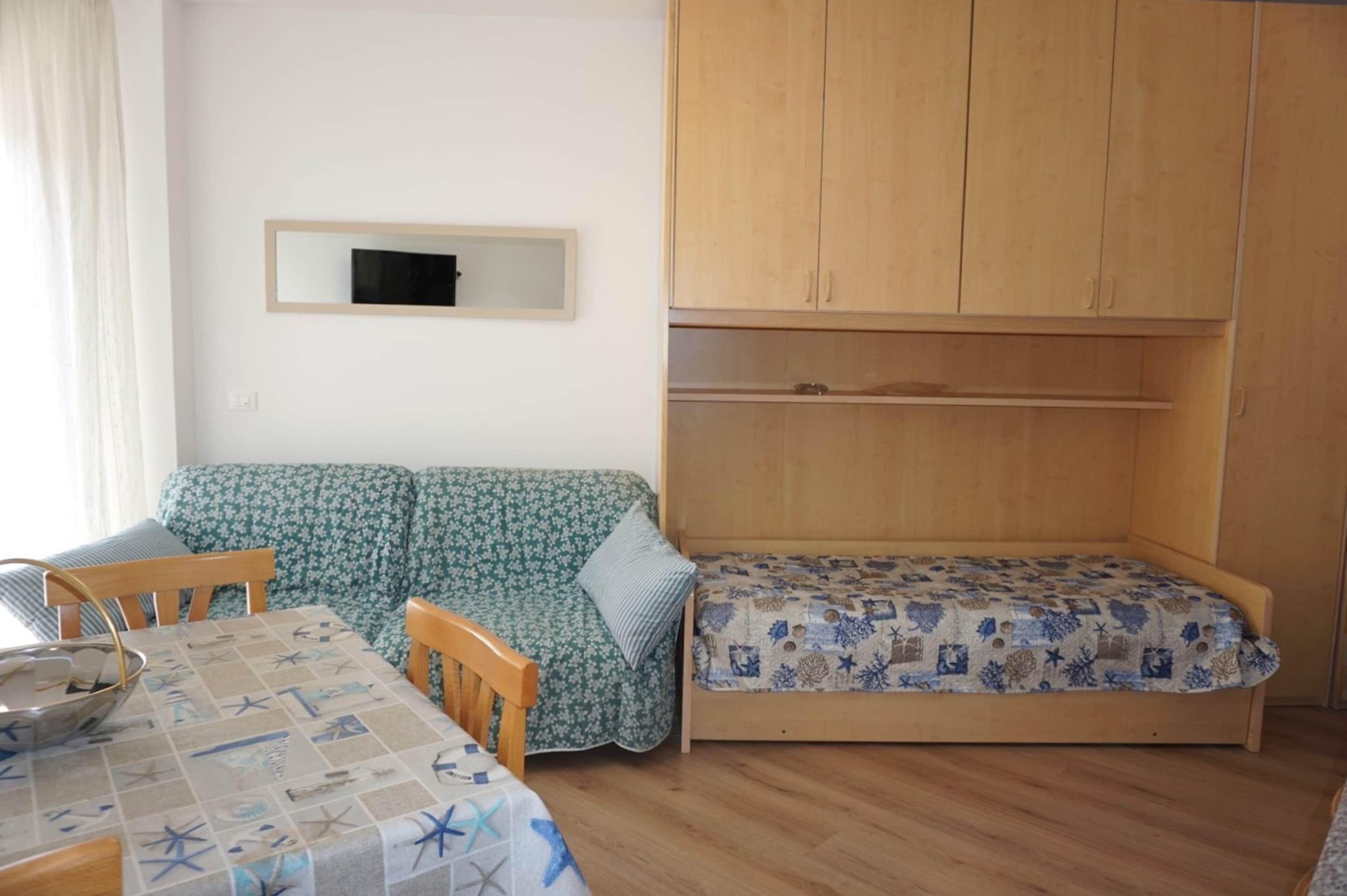 A room with a sofa, a bed, a table with a patterned tablecloth, and a wooden wardrobe.