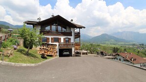 A two-story building with a balcony, a car parked in the driveway, and a mountainous landscape in the background. - Belvilla by OYO Corno Nero (Ville di Fiemme)