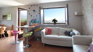 A living room with a large window, a sofa, a dining table, and colorful chairs. - Belvilla by OYO Corno Nero (Ville di Fiemme)