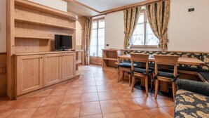 Un intérieur en bois avec un téléviseur posé sur une étagère, un coin repas avec des chaises et une table, et un canapé recouvert d’un tissu à motifs.