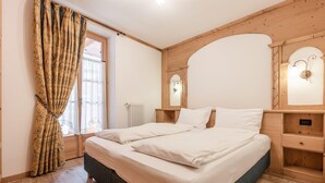 Une chambre à coucher avec une tête de lit en bois, un grand lit avec du linge de lit blanc, une fenêtre avec des rideaux et un luminaire fixé au mur.