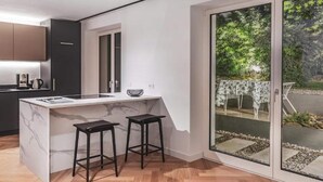 A modern kitchen with a marble island, two black stools, and a sliding glass door leading to an outdoor patio with a table and chairs.