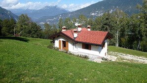 A small white house with a red roof and chimneys, surrounded by green grass and trees, with mountains in the background. - Belvilla by OYO Casera Pantonio (Fonzaso)