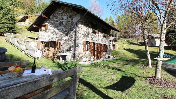 A stone building with a sloped roof, wooden shutters, and a wooden bench with a basket of fruit and a bottle. - Belvilla by OYO Baita Silana (Pieve Tesino)