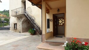 A building with a stone staircase leading to a balcony, a wooden door with a decorative glass panel, and a potted plant with red and white flowers in the foreground. - Family Retreat Near Garda (Ronzo-Chienis)