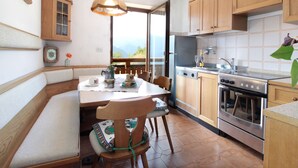 A kitchen with wooden cabinets, a dining table with chairs, a built-in oven, and a view through the window. - Large Apartment With Terraces in Val di Fiemme (Ville di Fiemme)