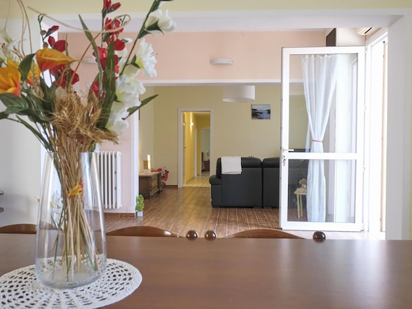 A dining area with a table, chairs, a vase with flowers, a radiator, a sofa, and a view through an open door.