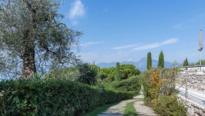 Un chemin de pierre traverse un jardin luxuriant, où poussent différentes plantes et arbres, et est bordé d’un mur de pierre et d’une rambarde en métal.