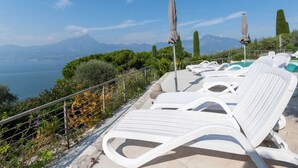 Un espace aménagé au bord de la piscine, avec des chaises longues blanches et des parasols, offrant une vue sur un plan d’eau et des montagnes.