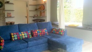 A blue sofa with colorful floral cushions, a glass coffee table, and shelves with various items including a guitar. - Belvilla by OYO Michael's House (Lesa)