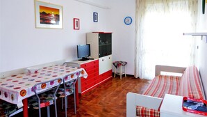 Ein Zimmer mit Esstisch, Stühlen, Fernseher, Schrank, einer Uhr und einem Fenster mit Vorhängen.