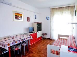 A room with a dining table, chairs, a television, a cabinet, a clock, and a window with curtains. - Holiday Home in Caorle Near Spiaggia Tartaruga (Caorle)