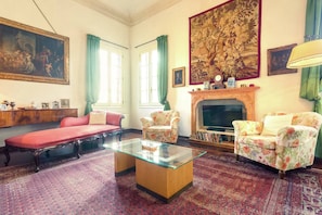 A room with a glass-top coffee table, two floral armchairs, a red sofa, a fireplace, and a large tapestry. - Villa in Venice With Private Pool & Park (Piombino Dese)