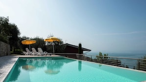 A rectangular swimming pool with clear blue water, surrounded by a wooden deck. There are two white lounge chairs and two yellow umbrellas. The pool is adjacent to a stone wall and overlooks a body of water. - Belvilla by OYO Gianni C Cinque Piao Due (Torri del Benaco)