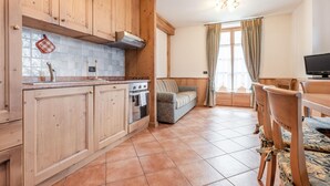 A kitchen with wooden cabinets, a tiled backsplash, and a built-in oven. There is a dining area with a wooden table and chairs, and a sofa in the background. - Belvilla by OYO Maso Margherita Quadrilocale (Commezzadura)