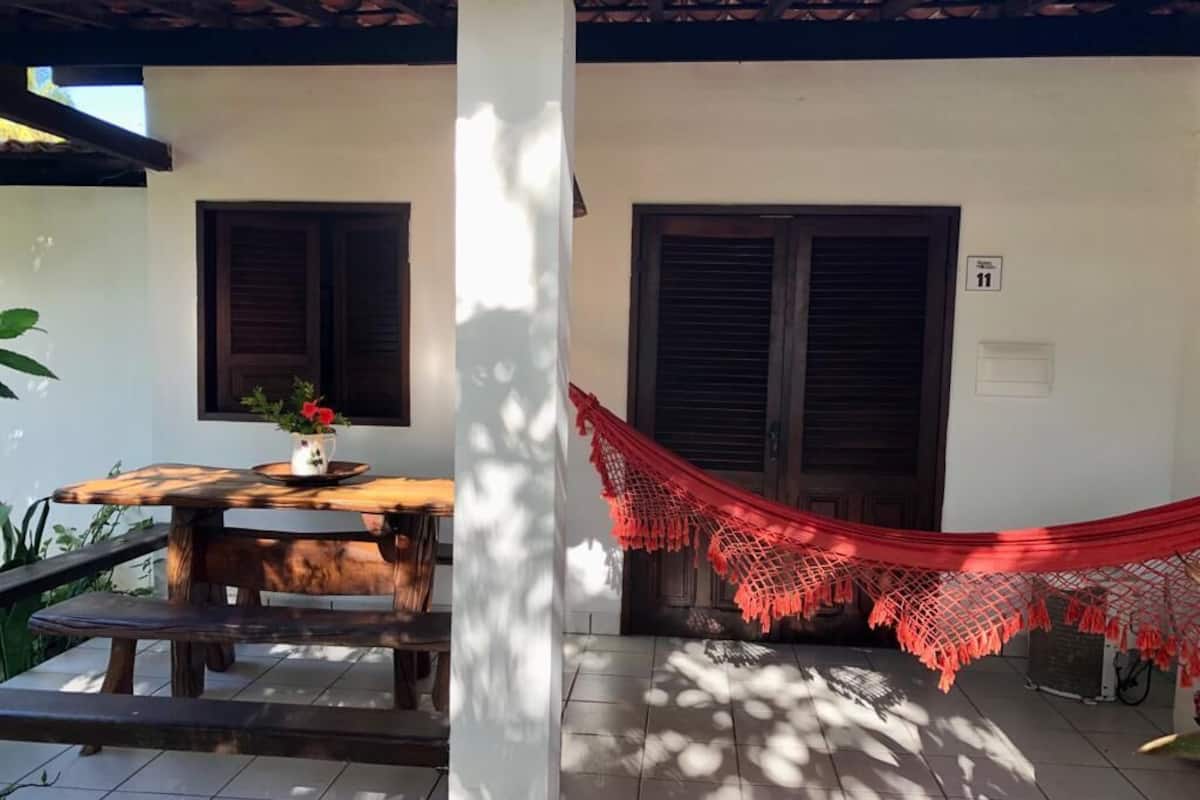 Um pátio com mesa e bancos de madeira, uma rede vermelha e um edifício branco com duas janelas e uma porta.
