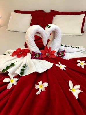Una cama con una colcha roja, almohadas blancas y una toalla dispuesta en forma de corazón.