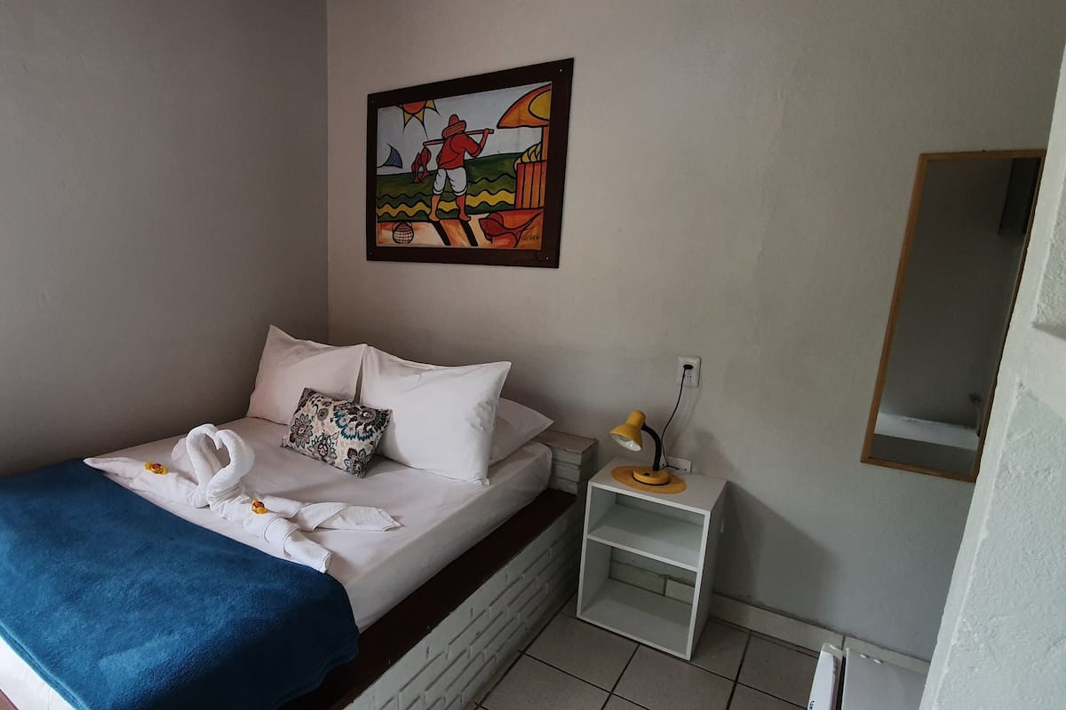 Uma cama bem arrumada com manta azul e almofadas brancas, uma mesinha de cabeceira com abajur e um quadro na parede.