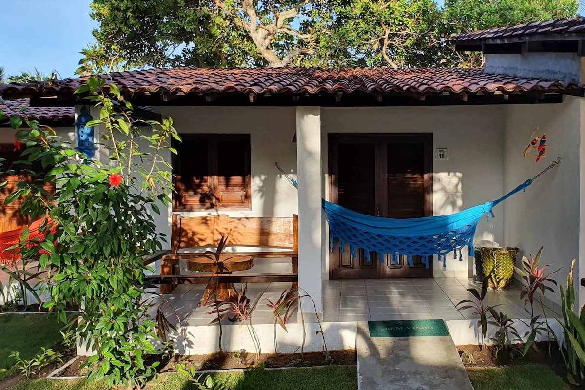 Uma casa com telhado de cerâmica, varanda com rede e pequeno jardim com diversas plantas.