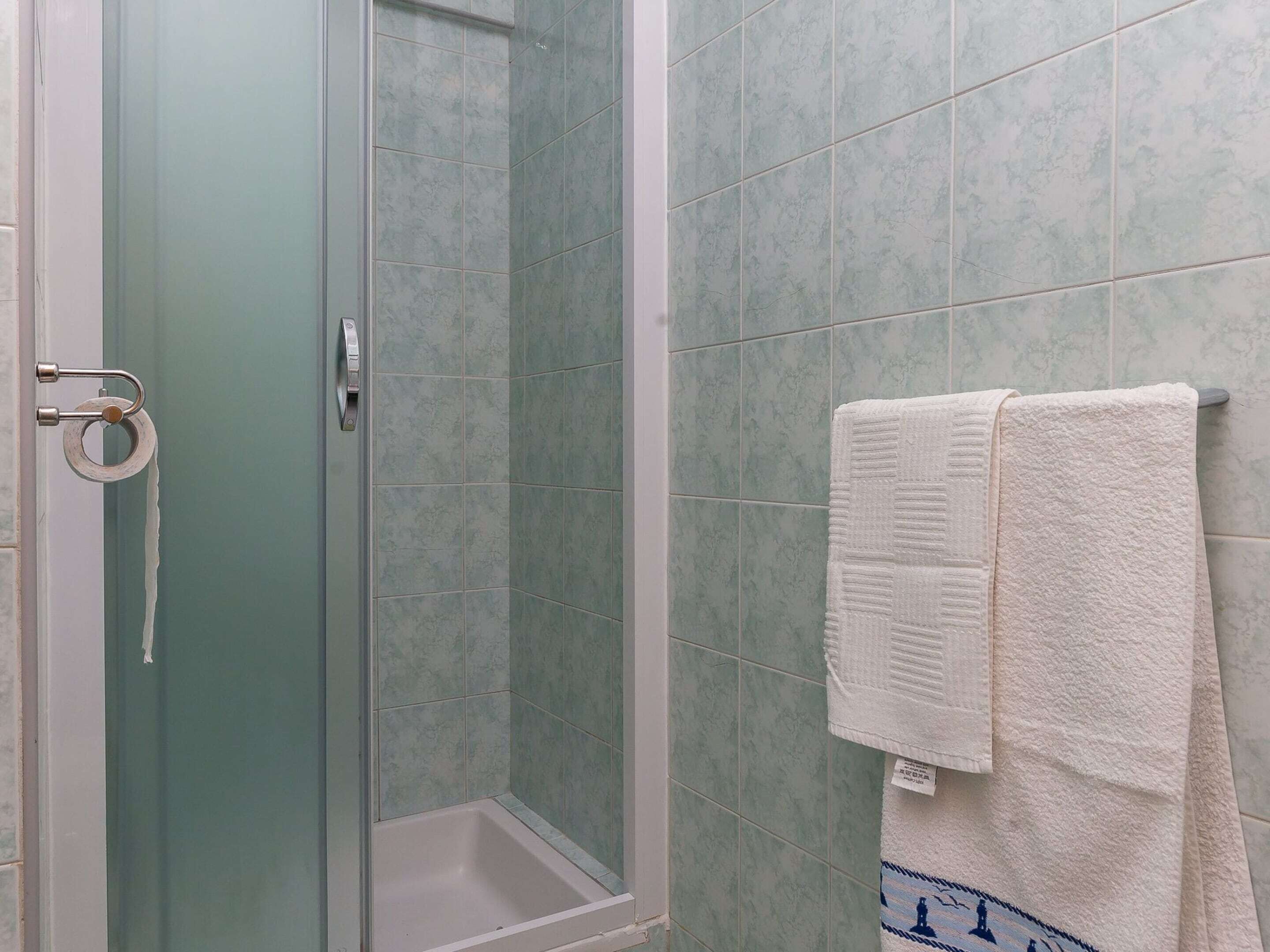 Ducha con puerta de vidrio, una toalla blanca colgada en un perchero y pared con azulejos verdes.