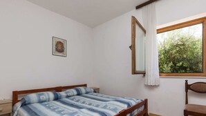 Ein Schlafzimmer mit einem hölzernen Bett, einem Nachttisch, einem Sessel, einem Fenster mit Blick auf Grünflächen und einem gerahmten Bild an der Wand.