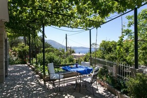 An outdoor patio with a table and chairs, surrounded by greenery and a view of the sea and mountains.