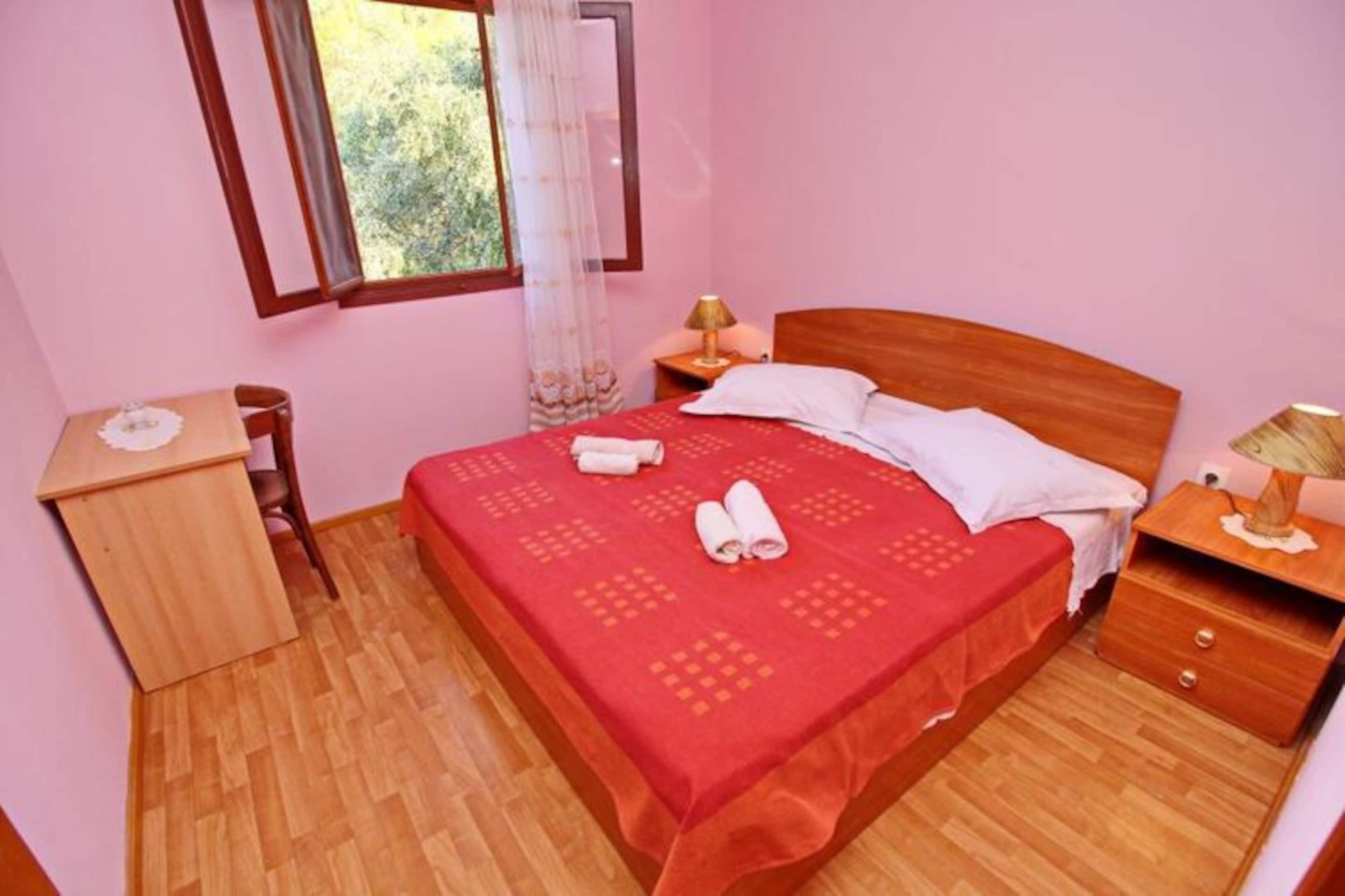 Quarto com cama de madeira, mesa de cabeceira, pequena escrivaninha e janela com vista para o verde.