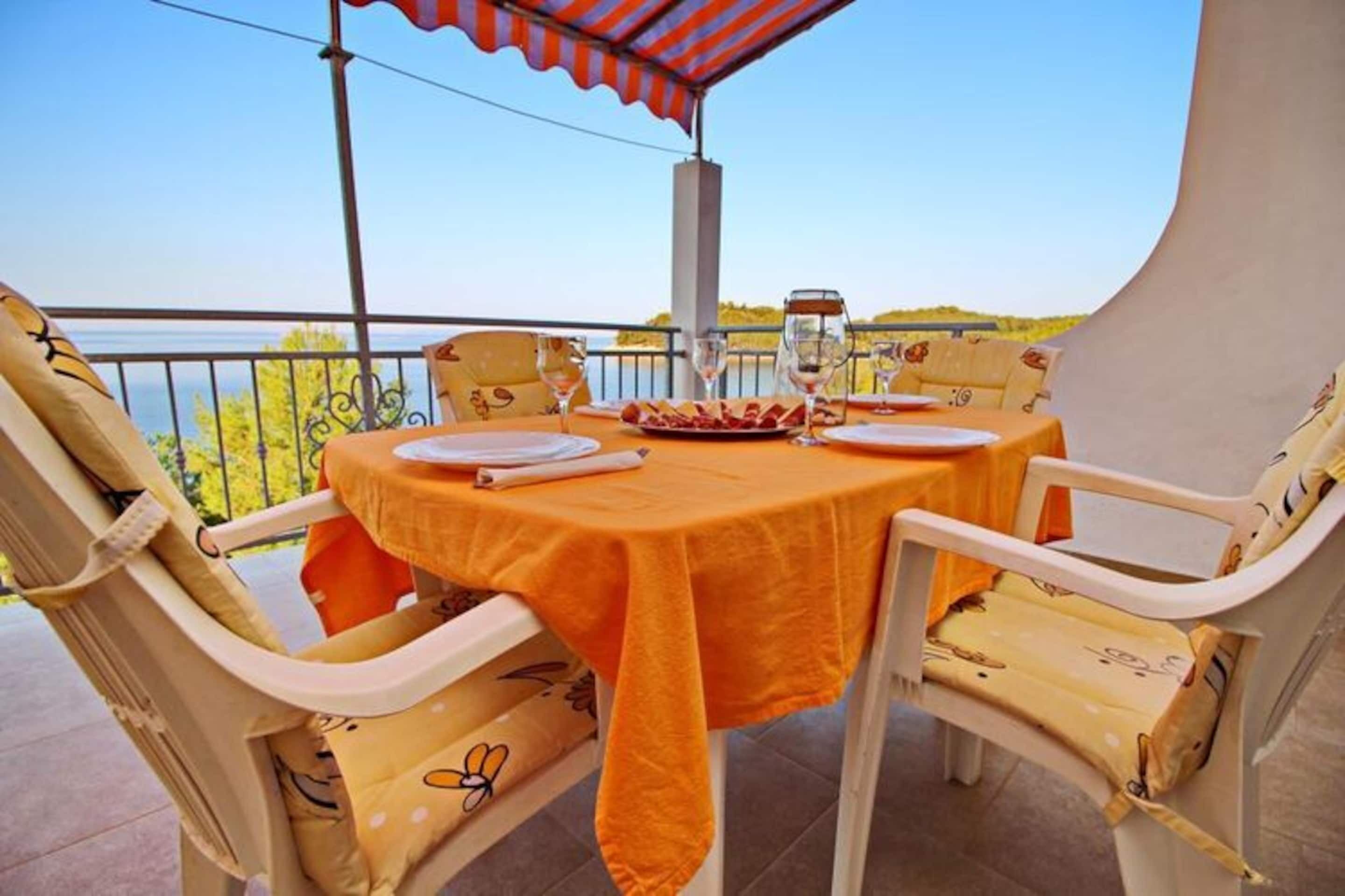 Una mesa redonda con mantel amarillo, cuatro sillas blancas con estampado floral y vista al mar.