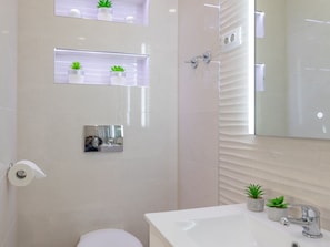 Ein modernes Badezimmer mit Toilette, Waschbecken und Spiegel. Über dem Spiegel befinden sich Blumentöpfe auf einem Regal.