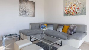 En moderne stue med en grå hjørnesofa, et glasbord og hvide lænestole. To abstrakte blomsterbilleder hænger på væggene.