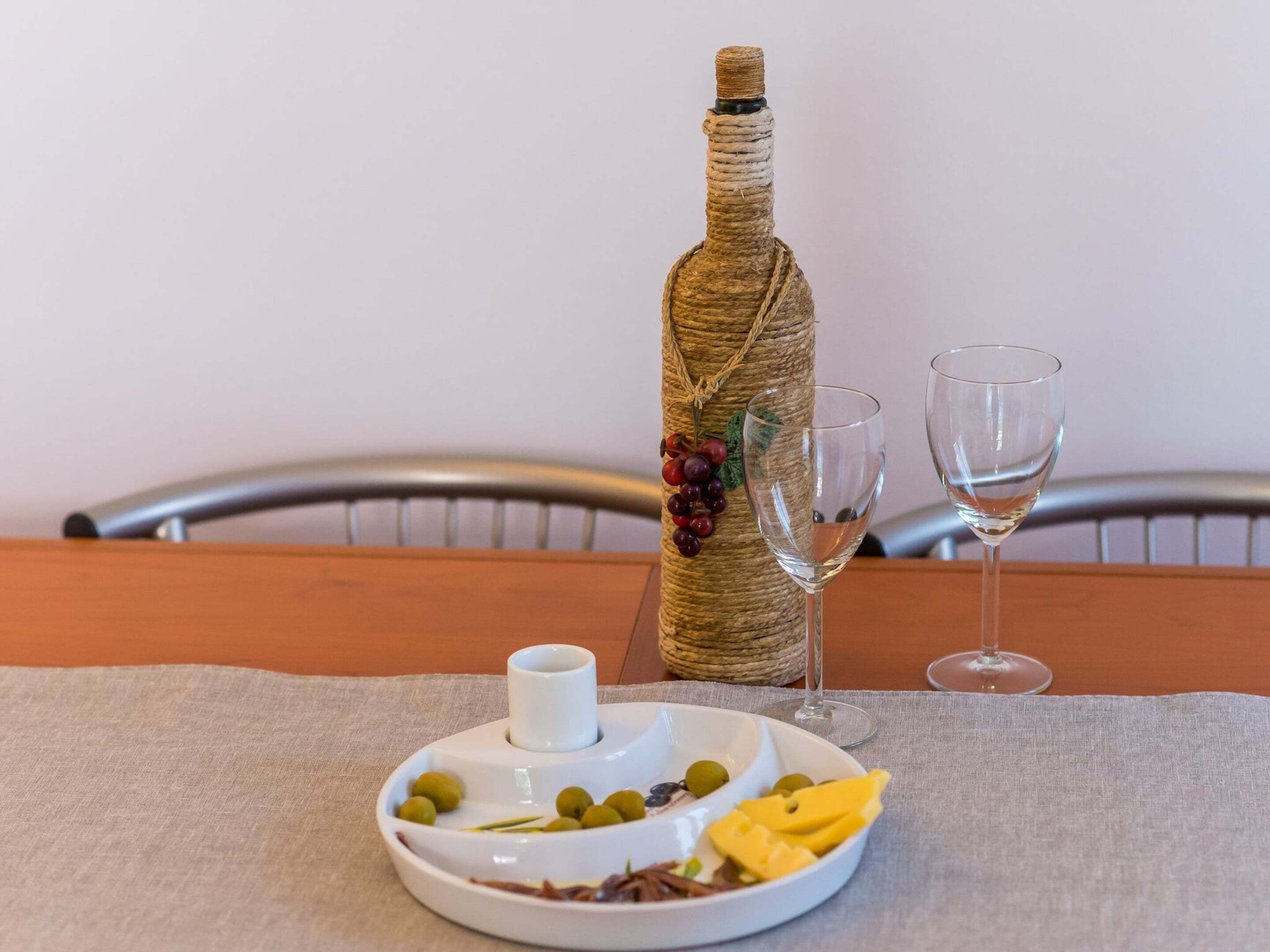 Une bouteille enveloppée de ficelle, deux verres à vin vides et une assiette avec des olives, des raisins et du fromage.