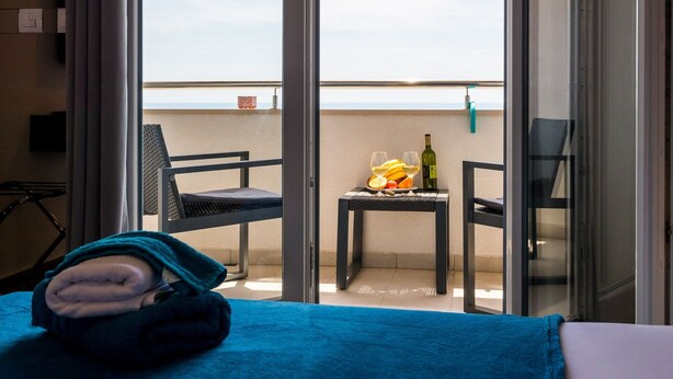 Un balcón con una mesa dispuesta con fruta, copas de vino y una botella. Hay una silla y una cama con cubrecolcha azul.