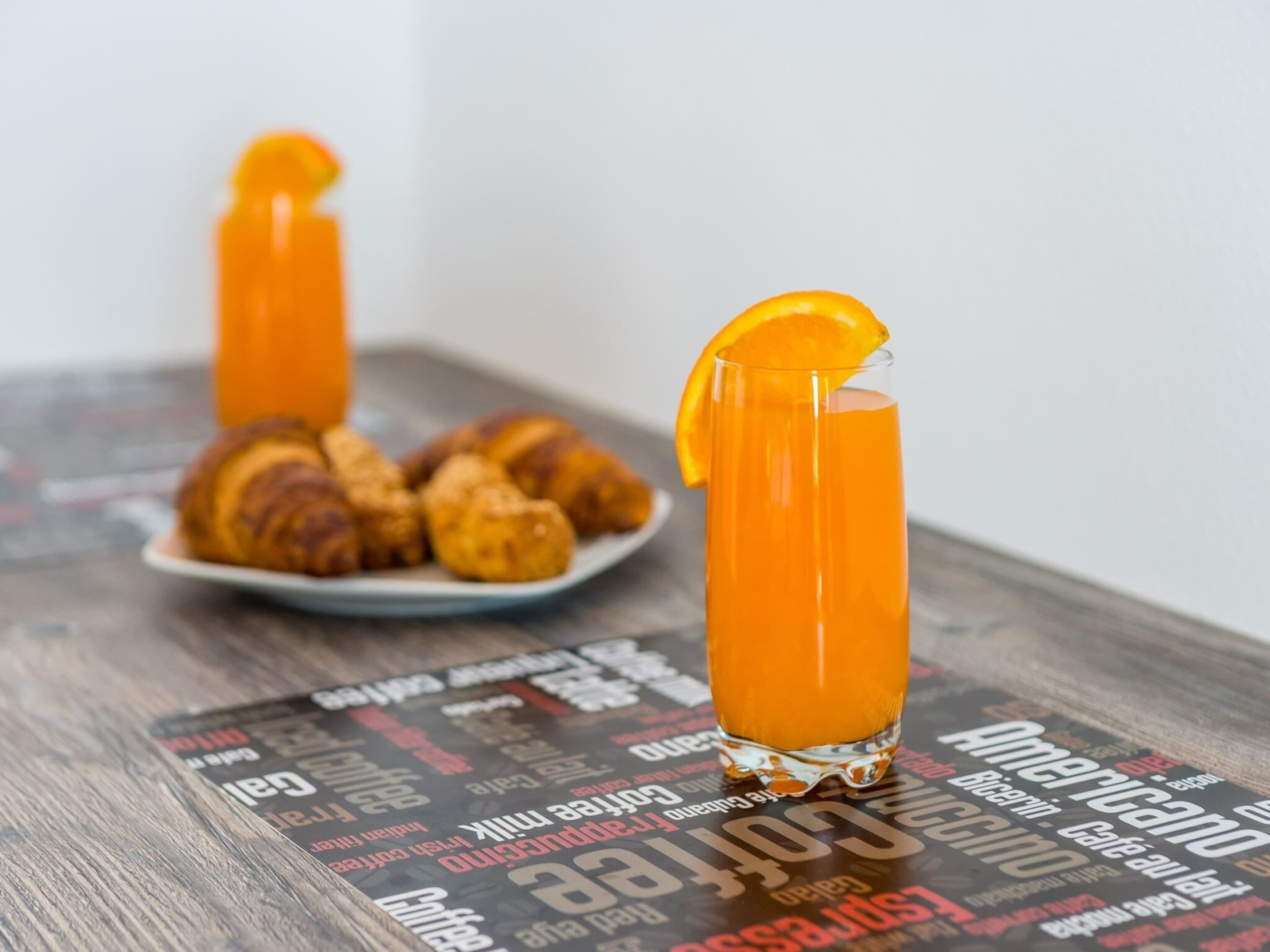 A glass of orange juice with a slice of orange on the rim, a plate of croissants, and a magazine with visible text.