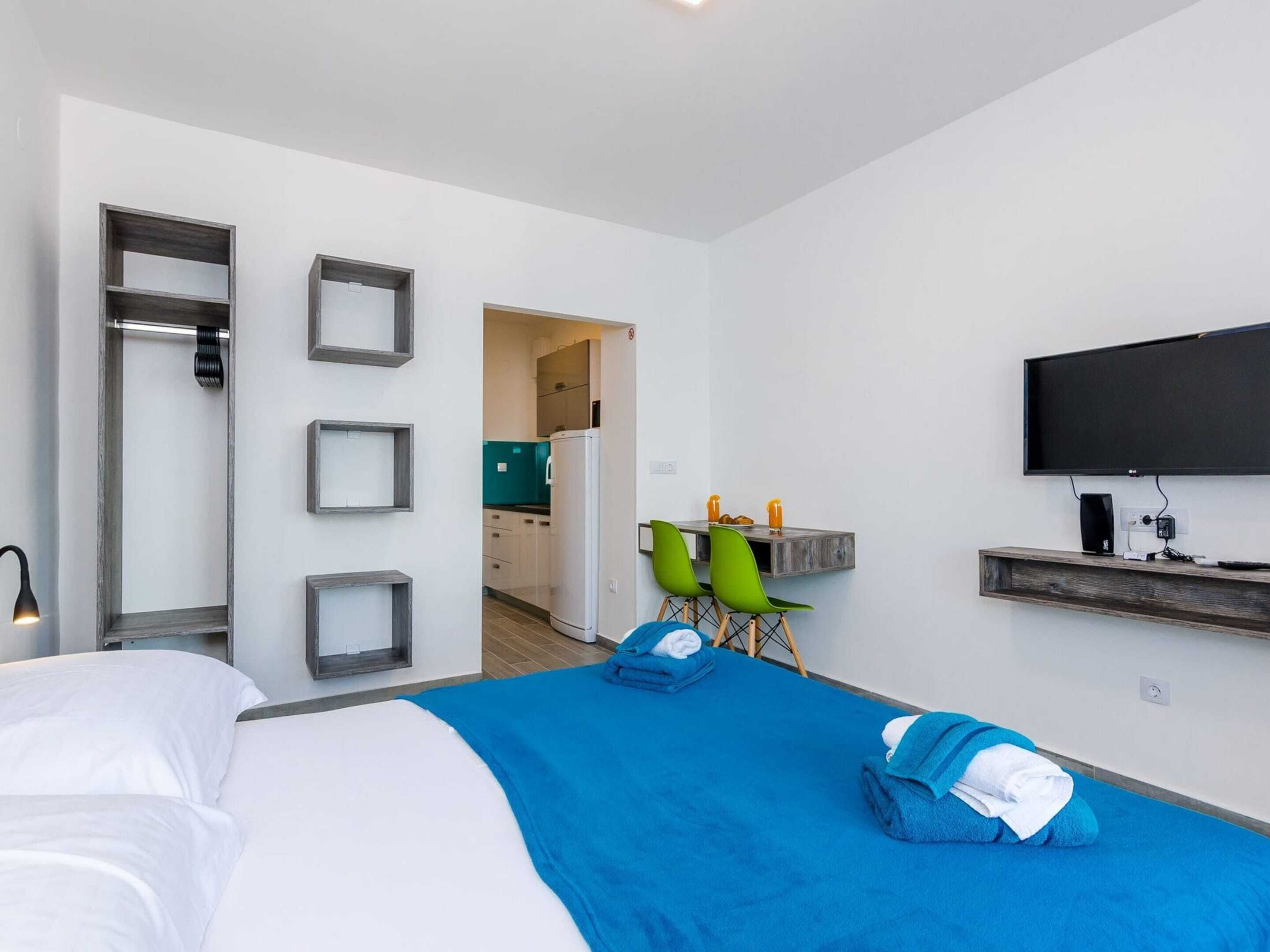 Une chambre d’hôtel avec un grand lit, un bureau avec une chaise, une télévision et, au fond, une petite cuisine.