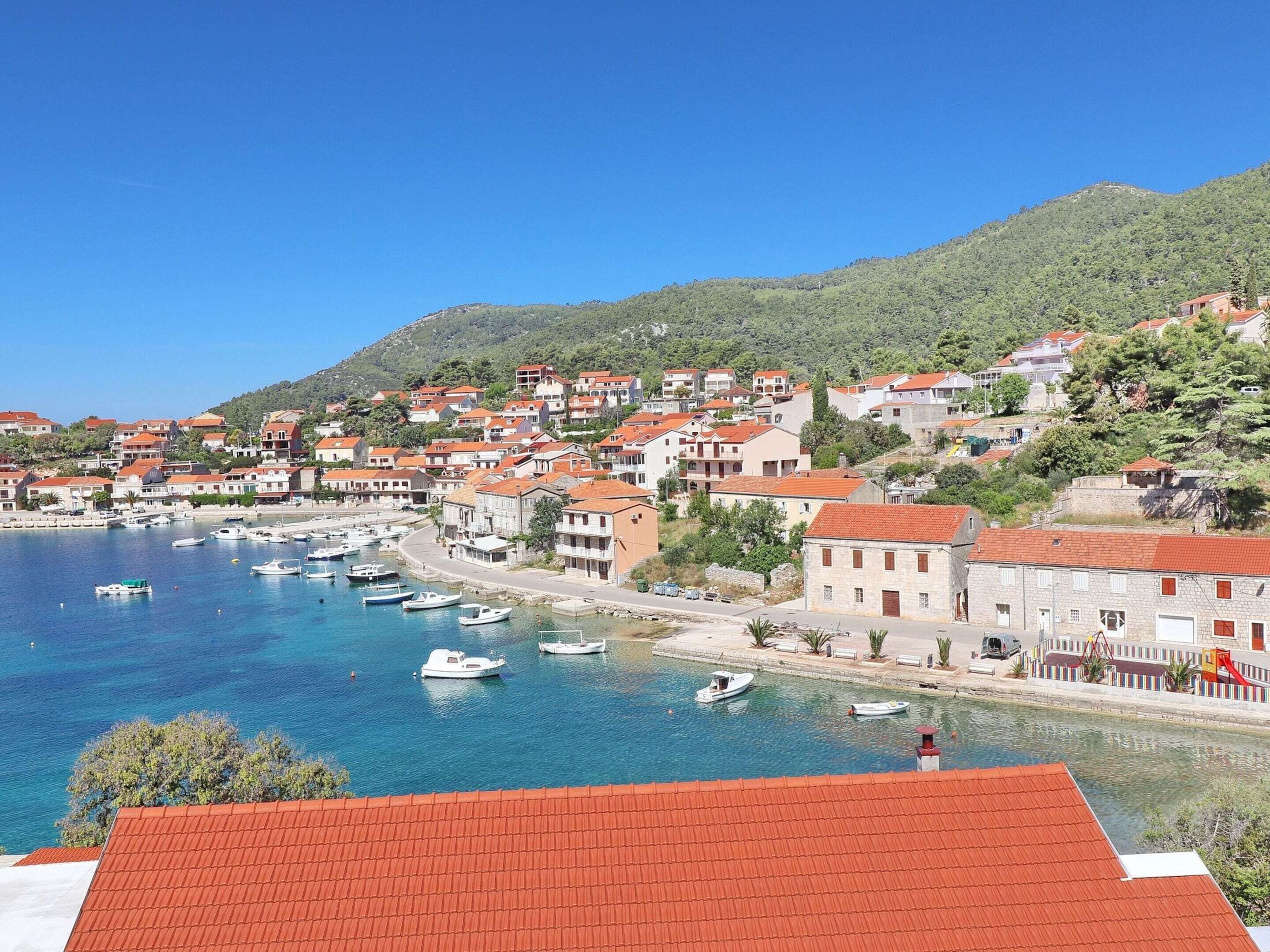 Una località costiera con edifici lungo il lungomare, barche ormeggiate nel porto turistico e una collina sullo sfondo.